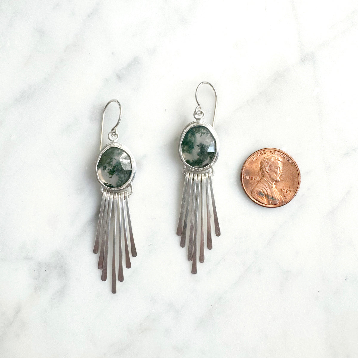 Silver fringe earrings with green stones on a white marble surface, next to a penny for scale.