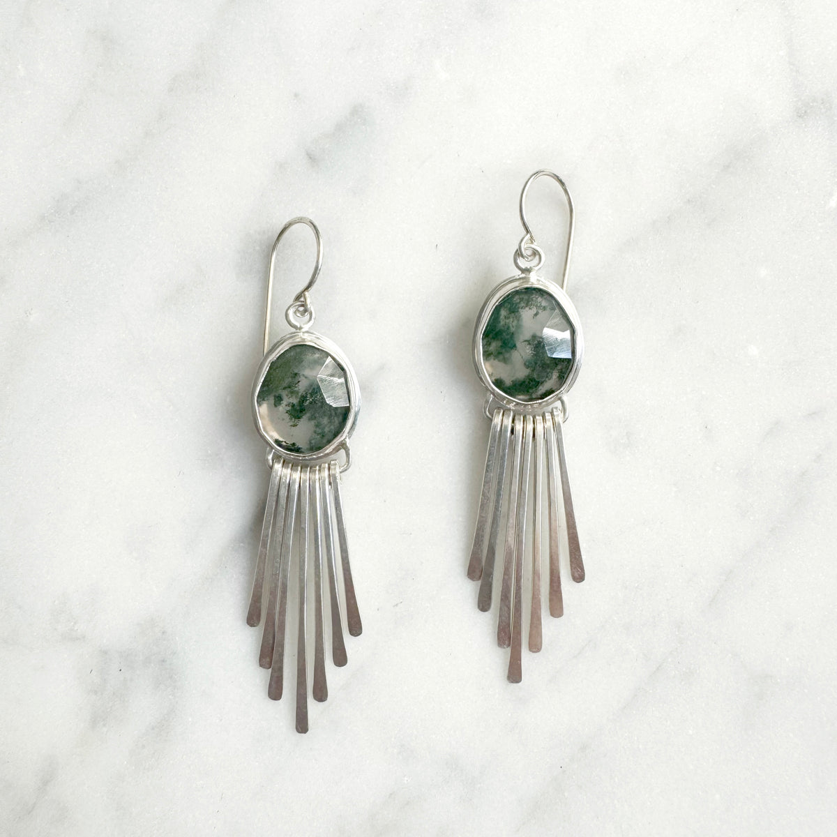 Silver fringe earrings with green stone accents on a white marble background