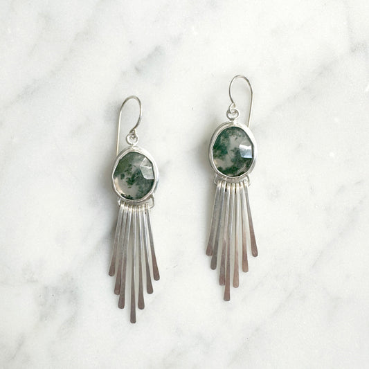 Silver fringe earrings with green stone accents on a white marble background