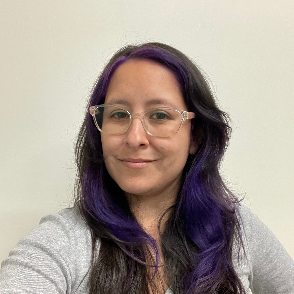 Person with purple hair wearing glasses and a gray sweater against a plain background