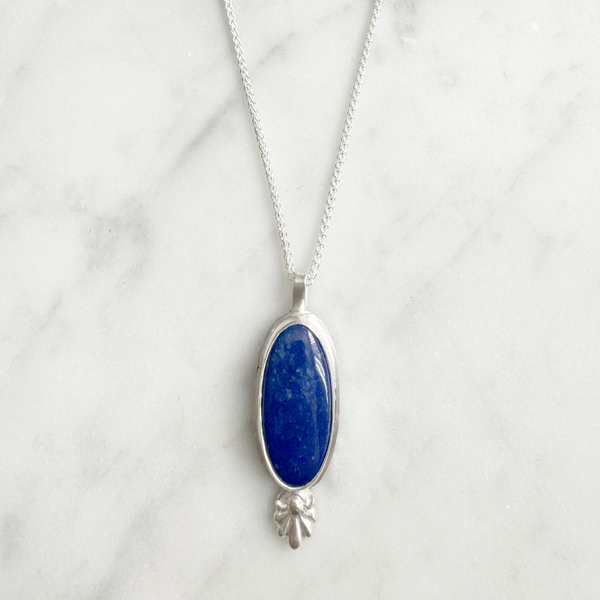Silver necklace with an oval blue lapis pendant with bottom ornament on a marble surface