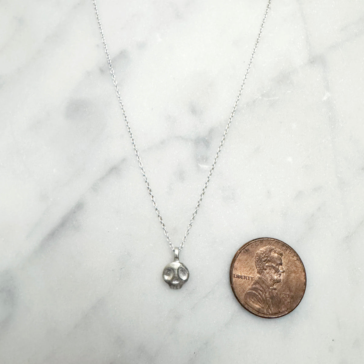 Silver necklace with a small skull pendant on a marble surface next to a penny for scale.